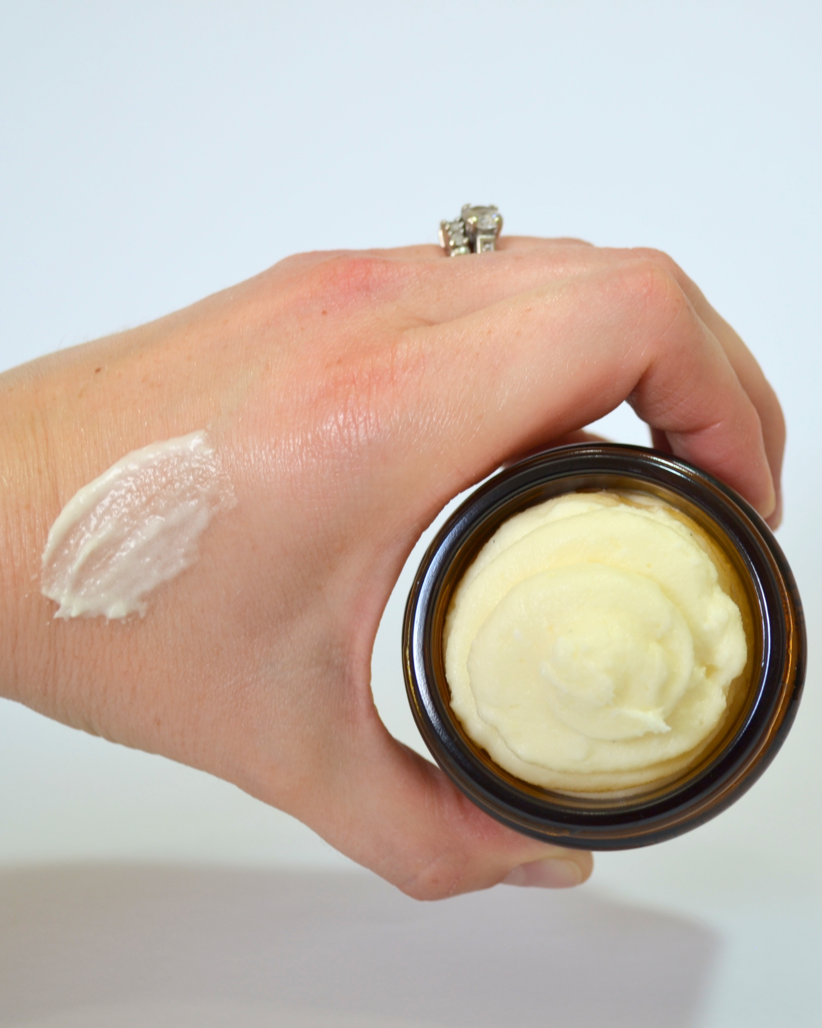 Hand holding a jar of cream with a visible sample of whipped tallow balm on skin, against a light background